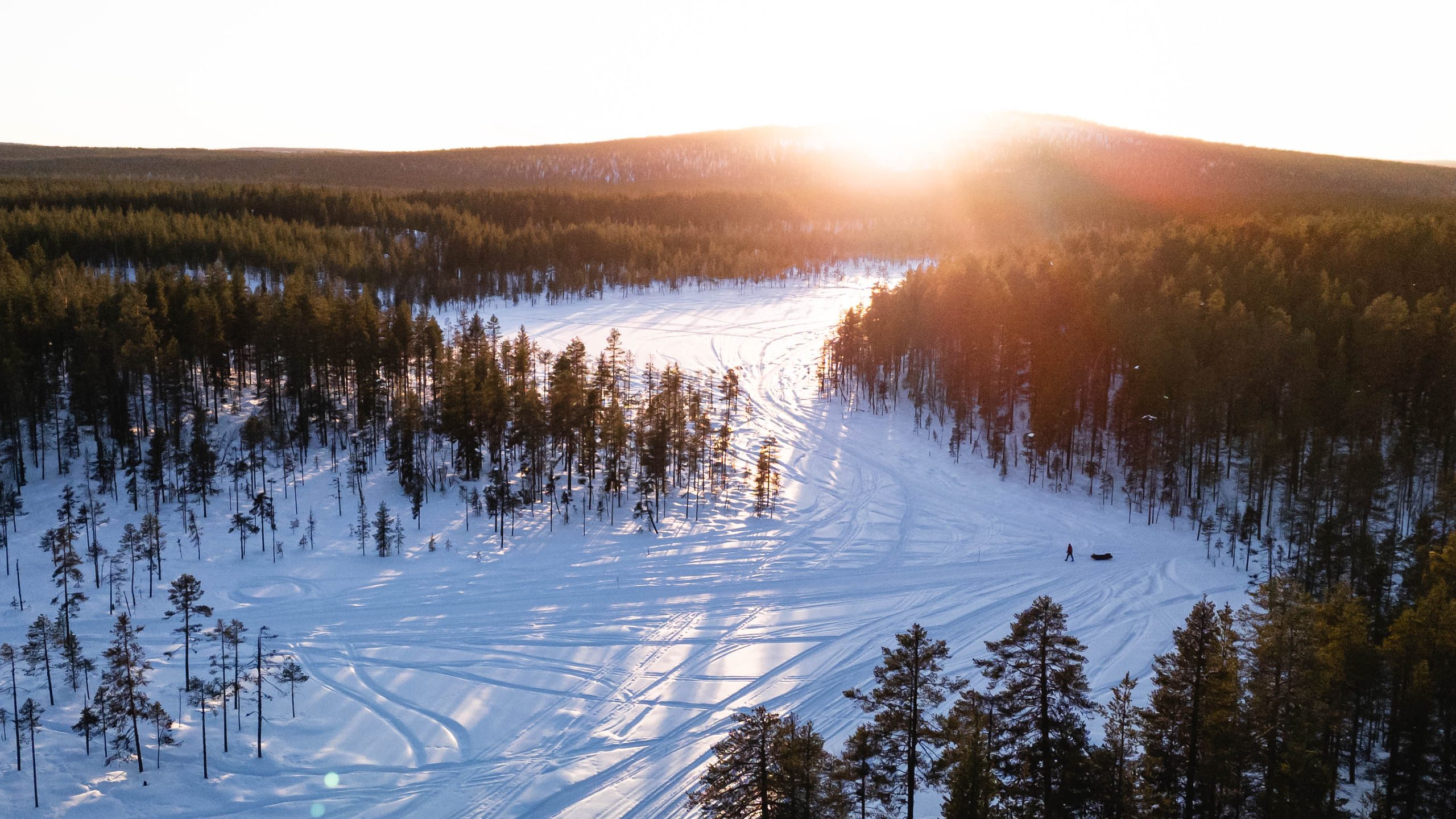 Lapland Arctic Ultra 2026, from the air at sunset: a lone figure pulling a sled across a snowy clearing in pine forest